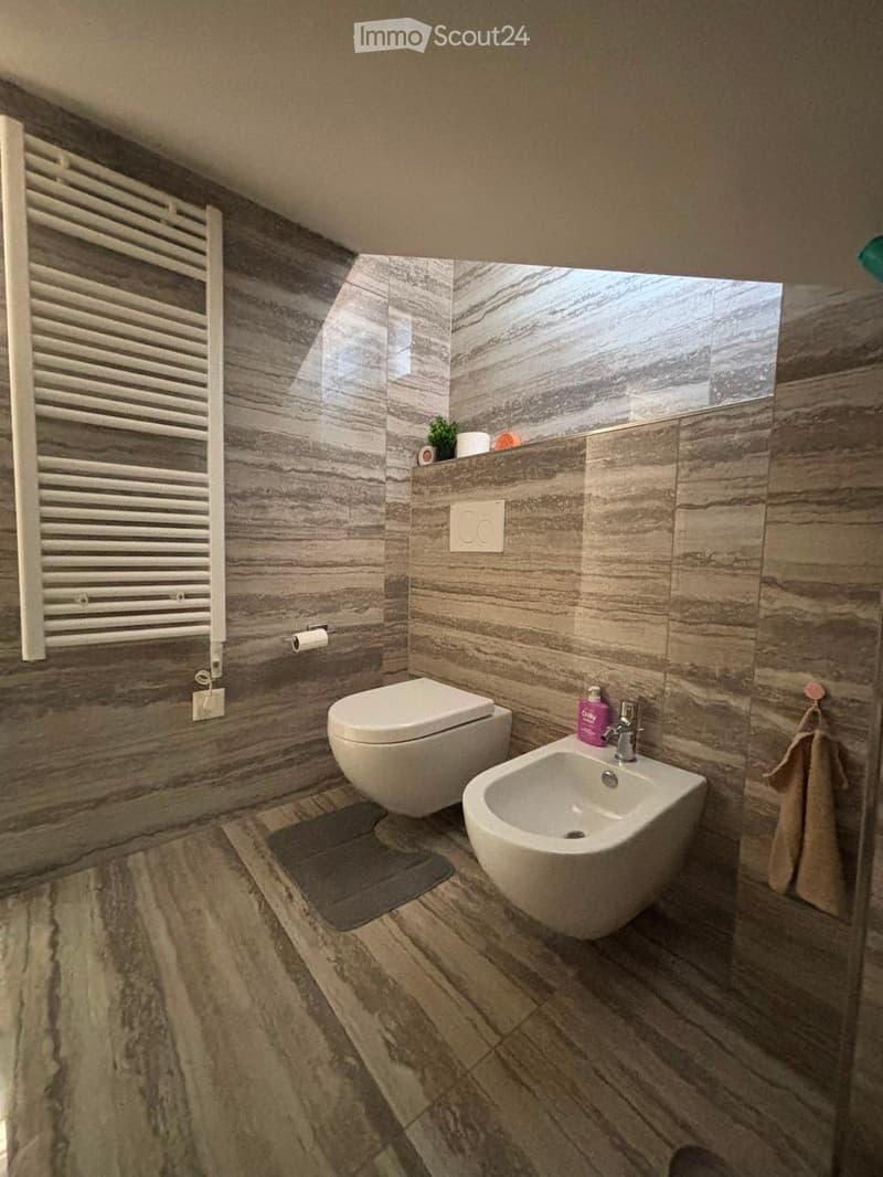 Modern bathroom with marble tiles, a toilet, a sink, a radiator, and a towel hanging on the wall.