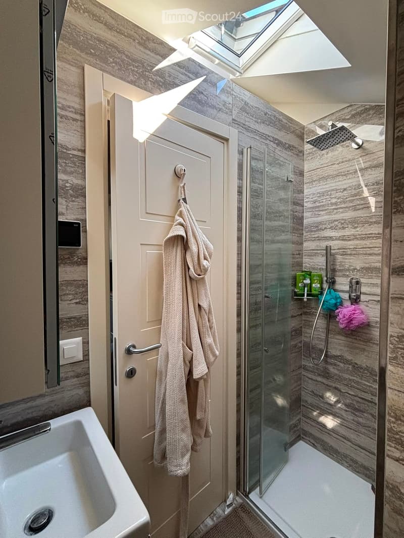 Modern bathroom with a bathtub, glass-enclosed shower, marble walls, and a skylight window above the shower area. A towel hangs on the door.