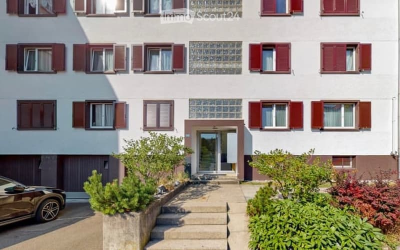 A two-story residential building with white walls, red shutters, and multiple windows. The entrance features a small staircase leading to a glass door, surrounded by small shrubs and plants.