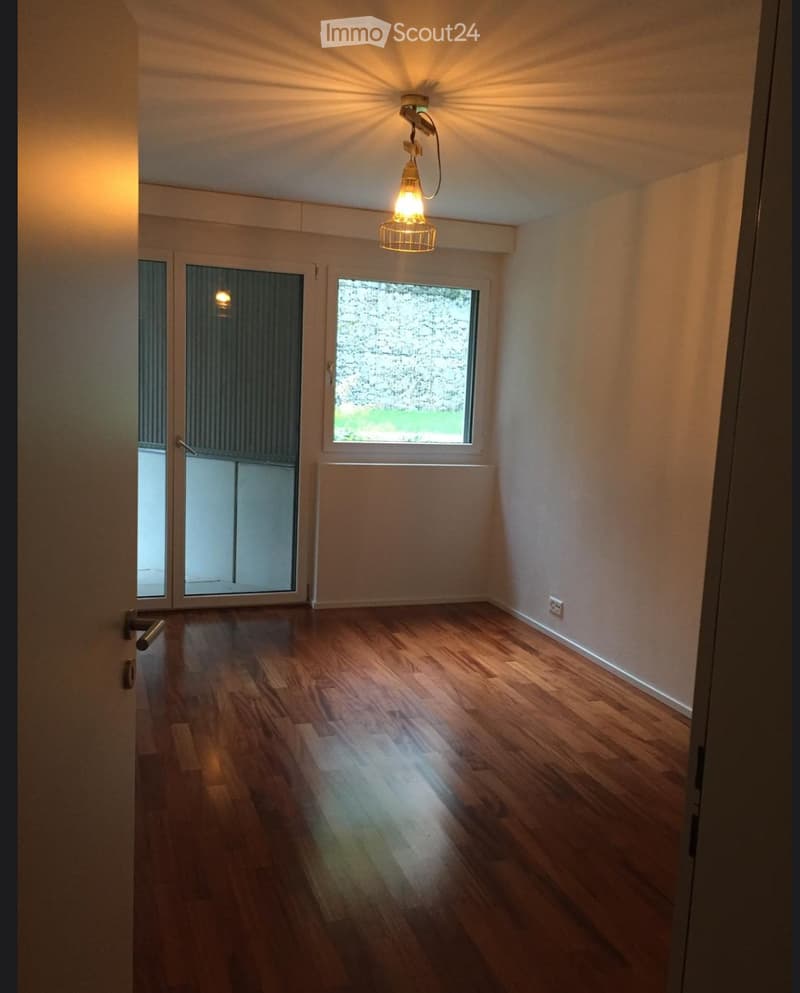 Empty room with wooden flooring, white walls, and large windows allowing natural light. Features a hanging light fixture and a door leading to another space.