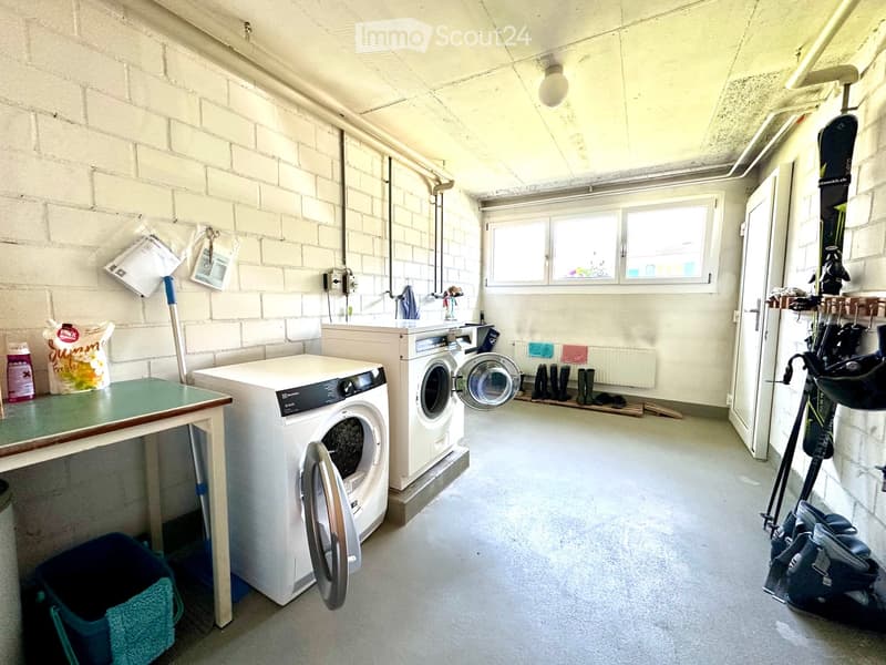 A small, well-lit laundry room with exposed brick walls, a washing machine, dryer, a small table, and various storage items including a backpack, snowboard, skis, and boots.