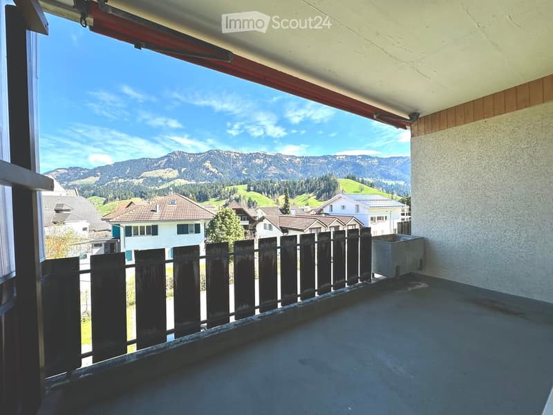 Balcony with wooden railing, overlooking a scenic mountain landscape and residential area.