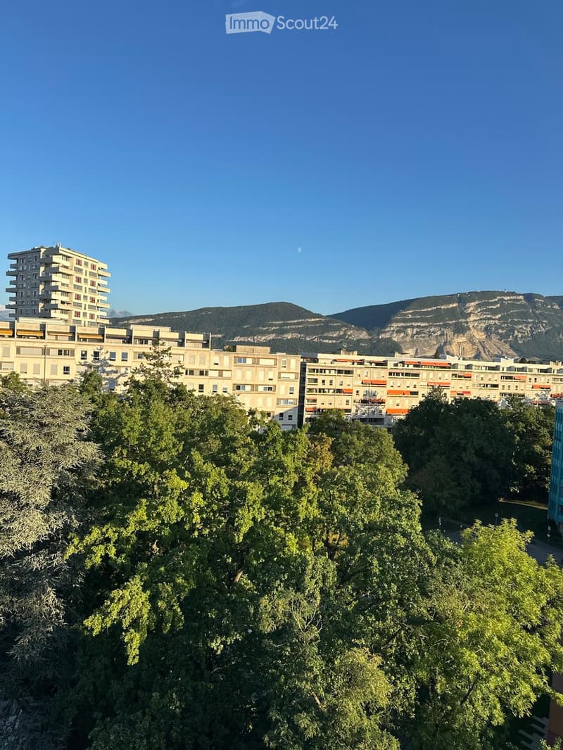 A view of a cityscape with multiple residential buildings, surrounded by lush green trees, with a mountain range in the background under a clear blue sky.
