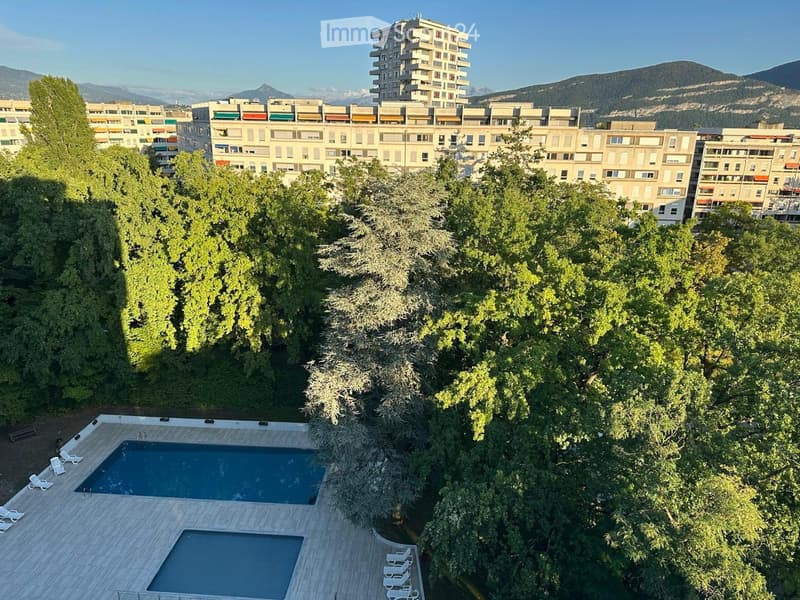 Aerial view of a rooftop garden with two swimming pools, surrounded by lush green trees and several lounge chairs, with apartment buildings and mountains in the background.