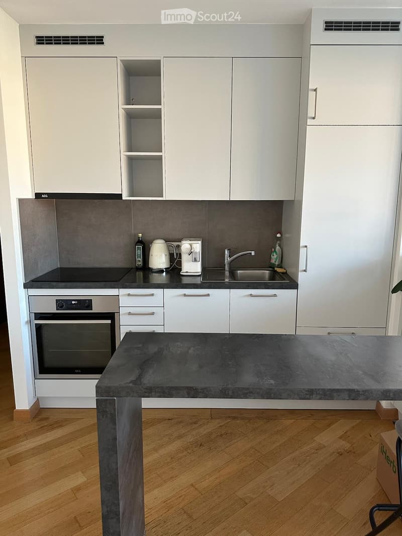 Modern kitchen with white cabinets, dark countertops, and wooden flooring. Features an oven, sink, coffee maker, kettle, and bottles on the counter.