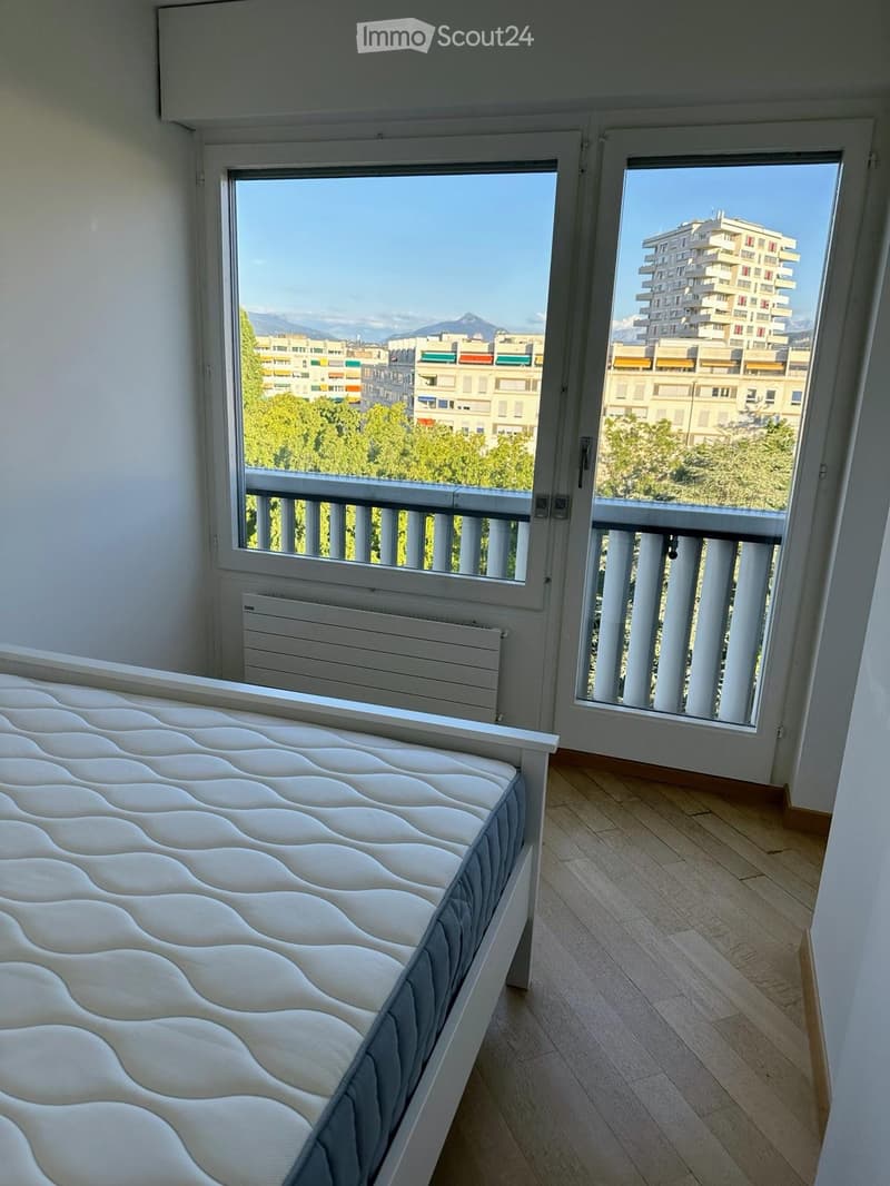 Bright bedroom with a white bed, wooden flooring, large windows offering a view of the city and mountains, and a radiator under one window.