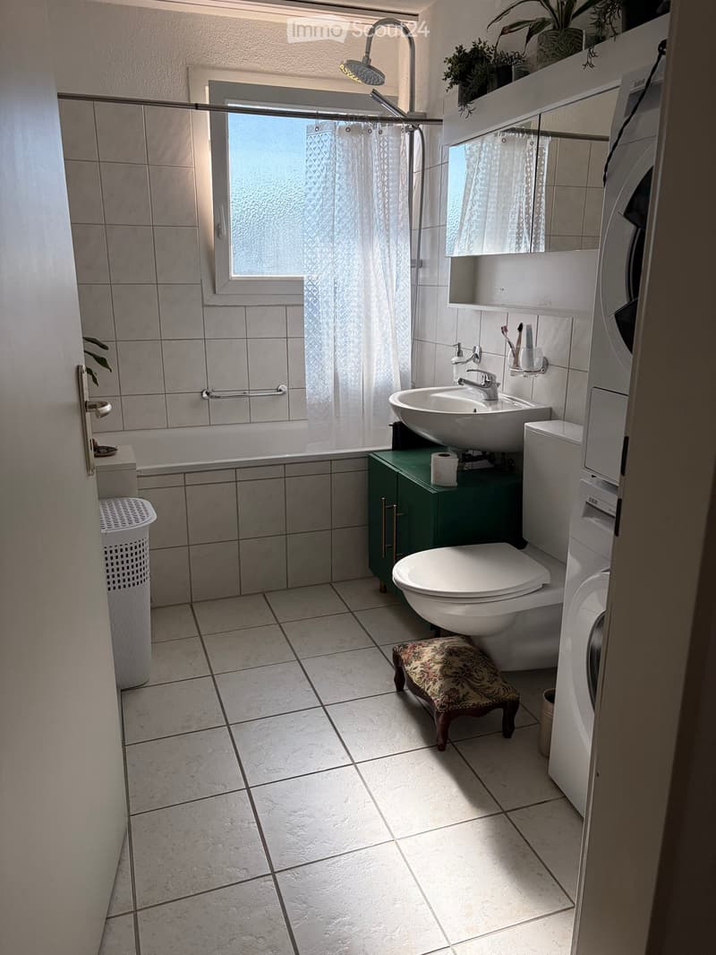 Modern bathroom with tiled walls and floor, featuring a bathtub, sink, toilet, and a small floral-patterned stool. Includes a washing machine/dryer unit, a mirror, and a window with a curtain.