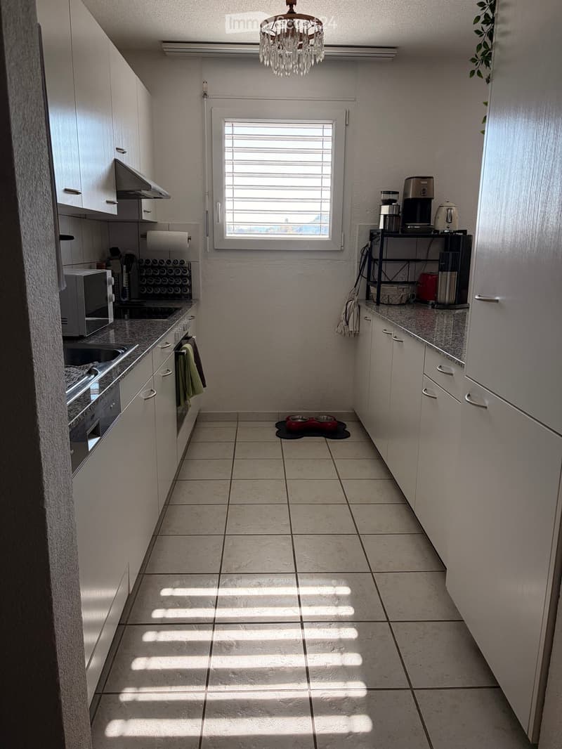 Modern kitchen with white cabinets, granite countertops, and tiled floor. Features a microwave, oven, sink, fridge, coffee maker, toaster, and a small chandelier.