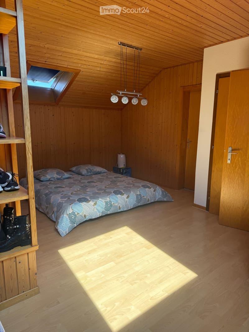 Cozy attic bedroom with wooden walls and ceiling, featuring a single bed, nightstand, pendant light, and a shelf with boots.