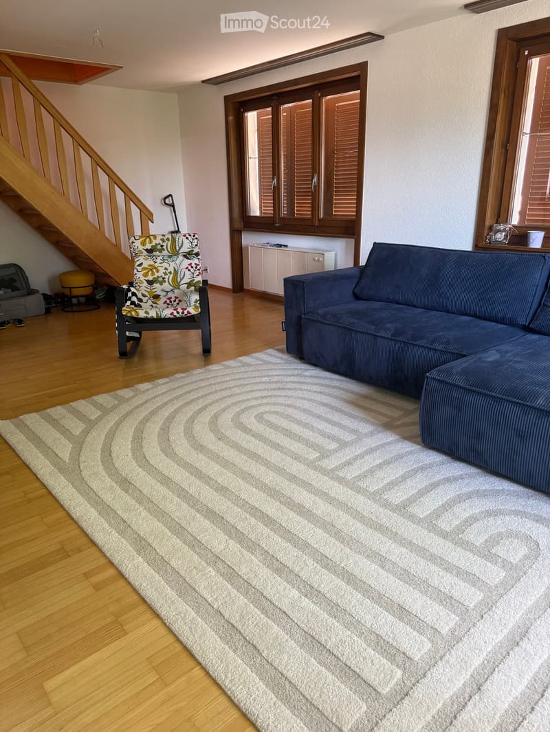 Living room with wooden flooring, a large sectional sofa, a patterned armchair, and a circular rug. A staircase leads upwards, and a window with shutters is visible.