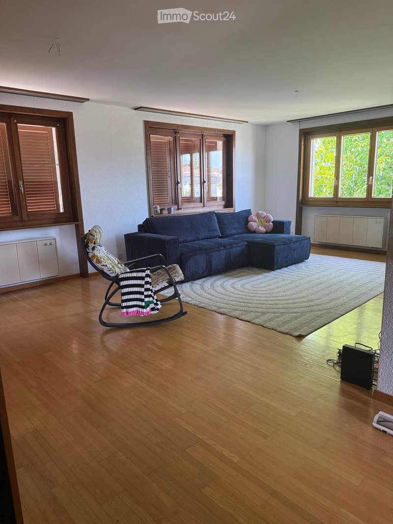 Spacious living room with wooden flooring, large windows, a sectional sofa, a rocking chair with a throw blanket, and a decorative pillow on the sofa. An area rug is placed in front of the sofa, and a speaker is visible on the floor near the window.