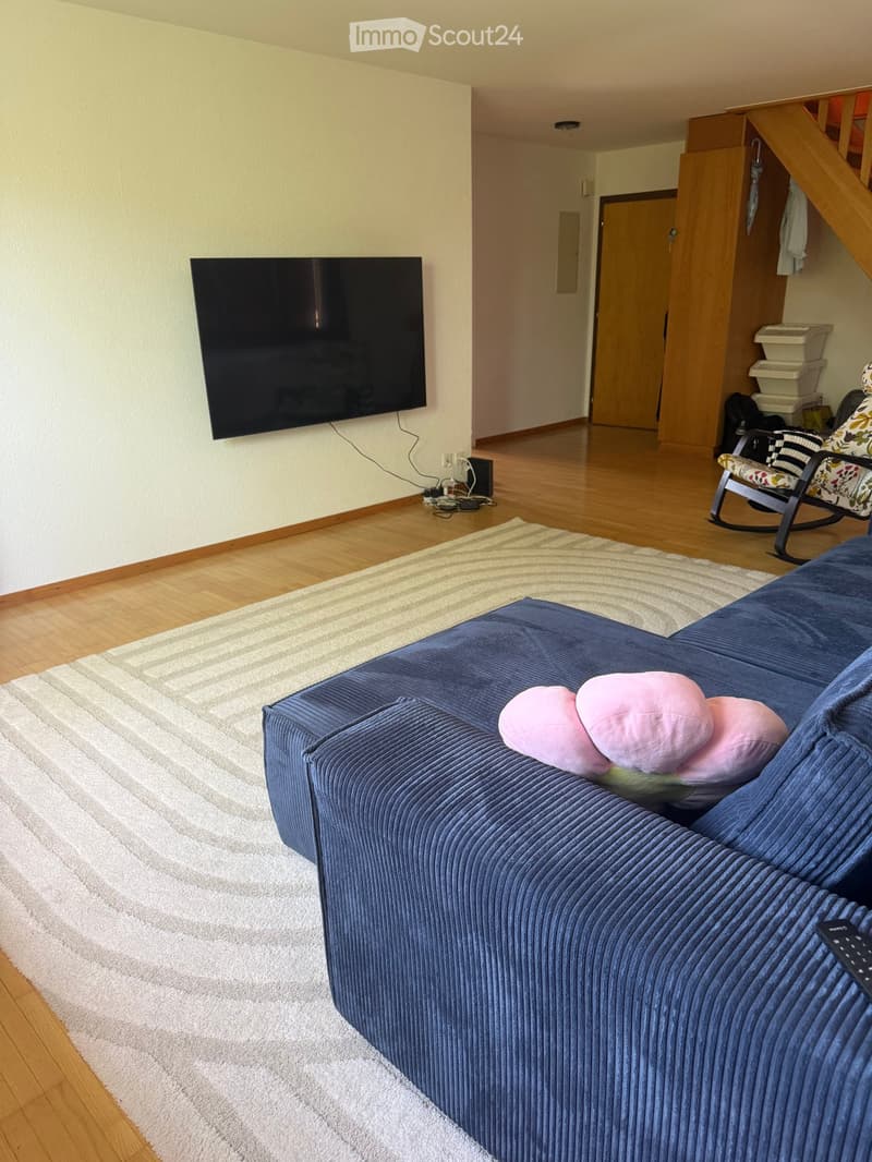 Living room with a blue sofa, pink pillows, a television mounted on the wall, a rocking chair, and a striped rug on a wooden floor.