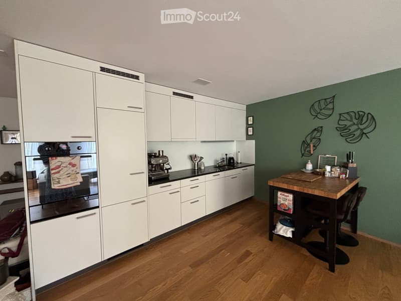 Modern kitchen with white cabinets, stainless steel appliances, wooden floor, island table with chairs, and various kitchen items on the counter.