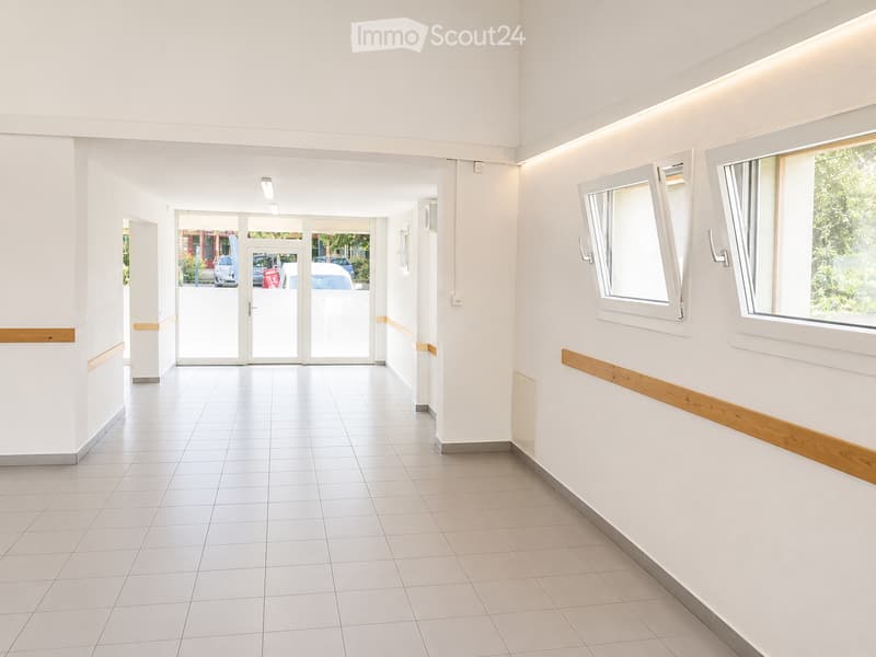 Bright, empty hallway with white tiled flooring, white walls, and large windows on one side. Features a double glass door at the far end leading outside, with wooden trim accents along the walls.