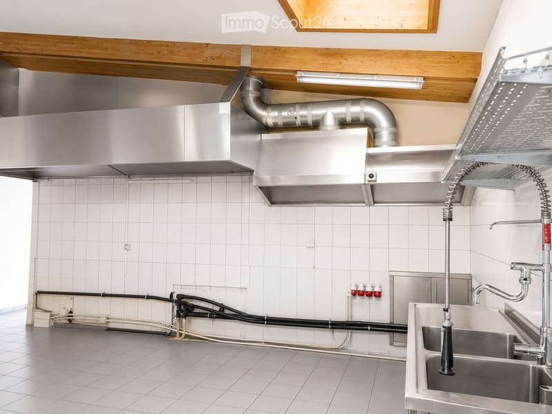 Commercial kitchen with stainless steel sink, faucet, range hood, and exposed pipes and wiring.