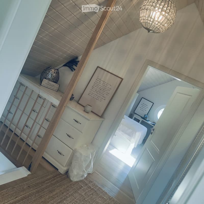 A well-lit hallway with wooden stairs, white walls, and a chandelier. A small shelf with decorative items is visible, and a doorway leads to another room.
