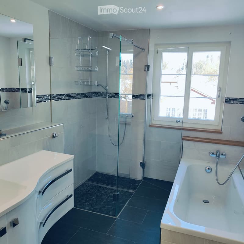 Modern bathroom with a bathtub, sink, mirror, and glass-enclosed shower. Features dark tiled floors and light-colored walls.