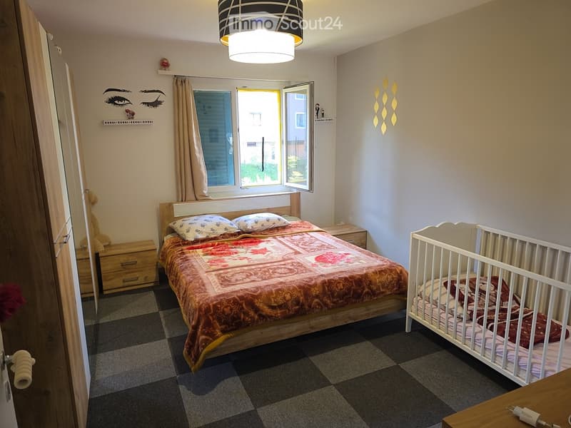 Cozy bedroom with a double bed, crib, nightstand, and decorative wall art. Features a large window with curtains and a modern pendant light.