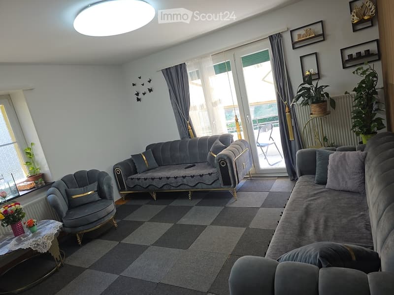 Living room with grey sofas, a small round table with a flower vase, a chair, and a radiator. The room features large windows with curtains and a view of the balcony.