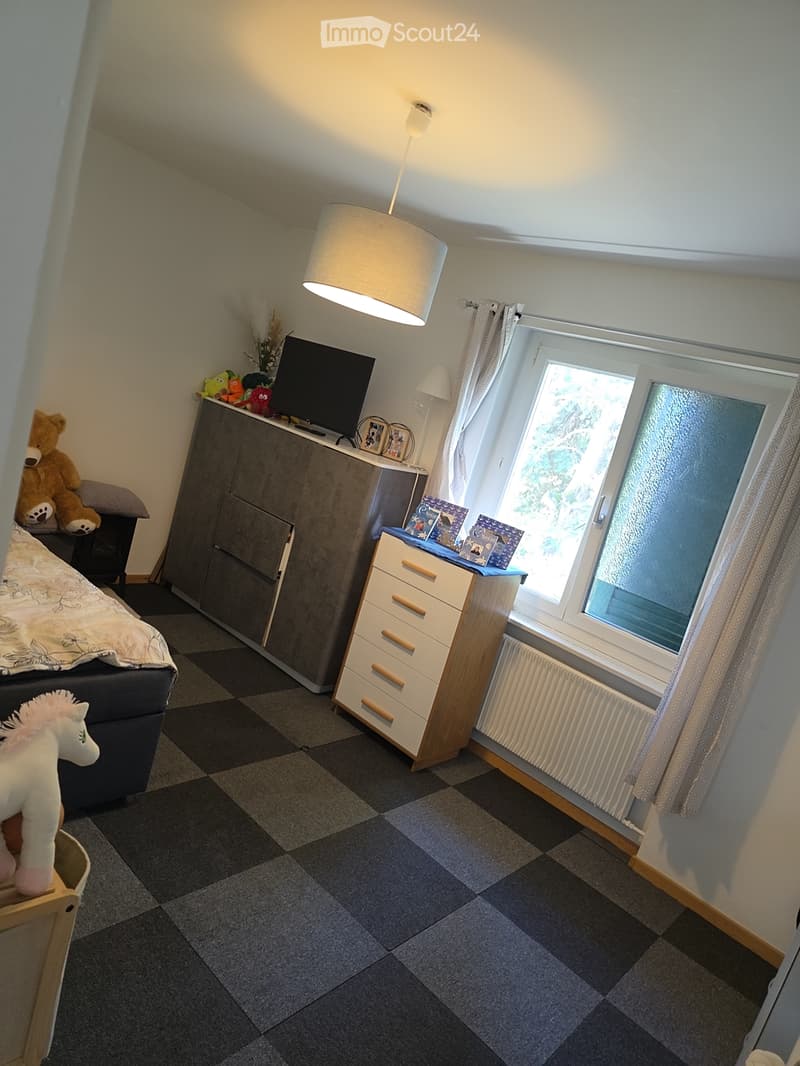 Cozy bedroom with a bed, nightstand, lamp, drawer unit, and a window with curtains. The room features a radiator under the window and a variety of toys on the nightstand.