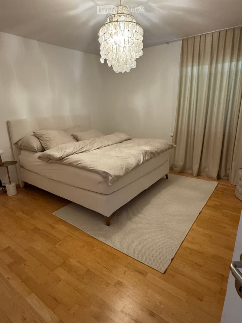A modern bedroom with a large bed, white bedding, a chandelier, and a nightstand. The room features wooden flooring and a white rug under the bed.