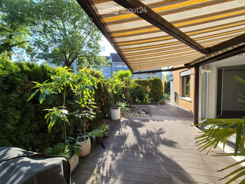 Wooden deck with potted plants, covered patio area with striped awning, lush greenery, and a view of neighboring houses.