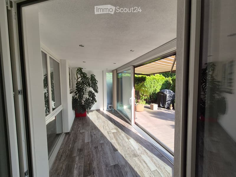 Bright hallway with wooden flooring, large windows, and sliding glass doors leading to an outdoor patio area with plants and a covered seating area.