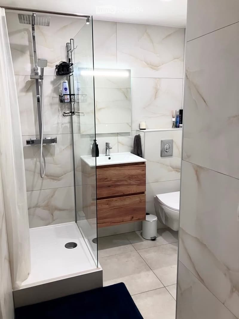 Modern bathroom with a glass-enclosed shower, wooden vanity cabinet, white sink, toilet, and tiled walls.