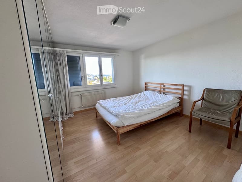 A small, well-lit bedroom with wooden flooring, a single bed with white bedding, a wooden chair, and a window with curtains offering a view of the outside.