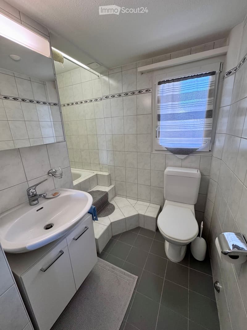 Modern bathroom with a white sink, toilet, bathtub, and mirror. Features tiled walls and a window with blinds.