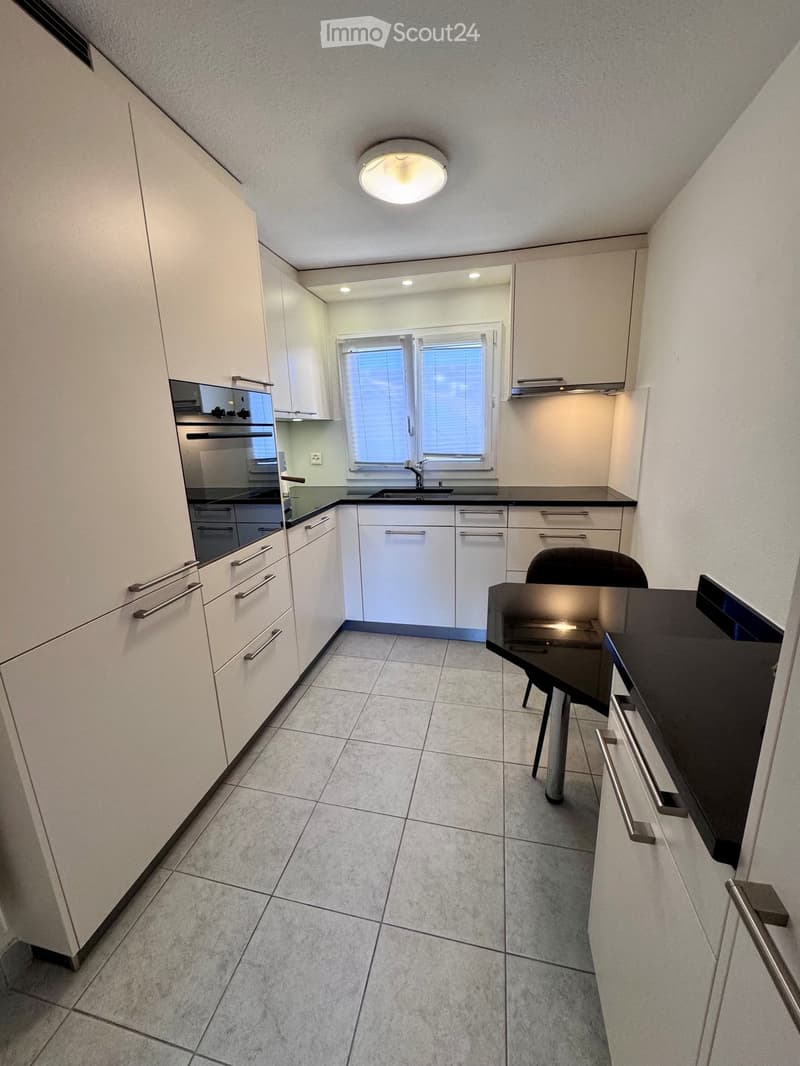 Modern kitchen with white cabinets, stainless steel appliances, tiled floor, and a small dining area with a black table and chair.