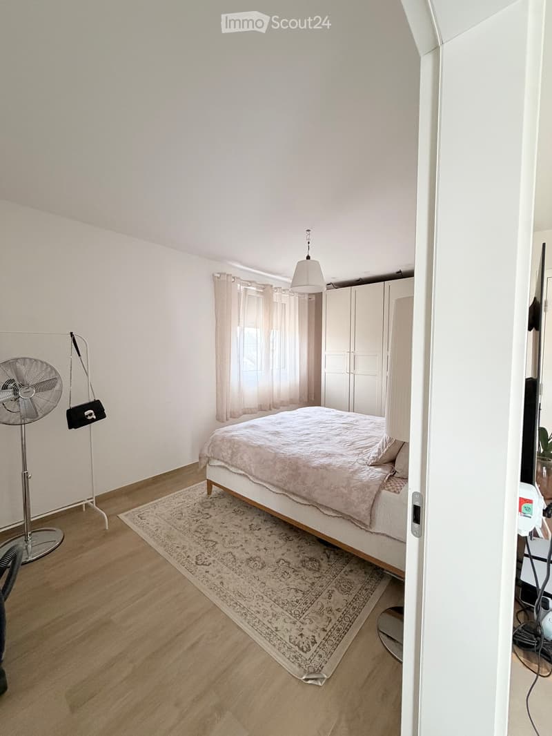 Bedroom with wooden flooring, a bed with a pink duvet, a fan, a wardrobe, and a curtained window.