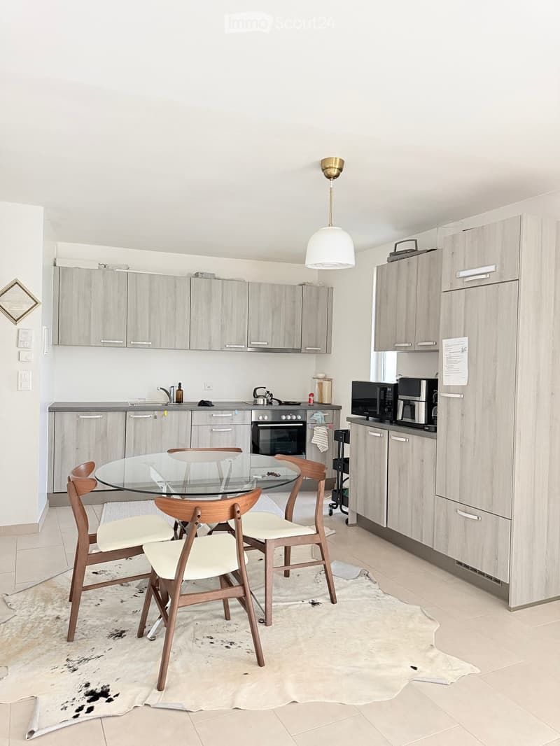 Modern kitchen with light wood cabinets, stainless steel appliances, a glass dining table with four wooden chairs, and a hanging pendant light.