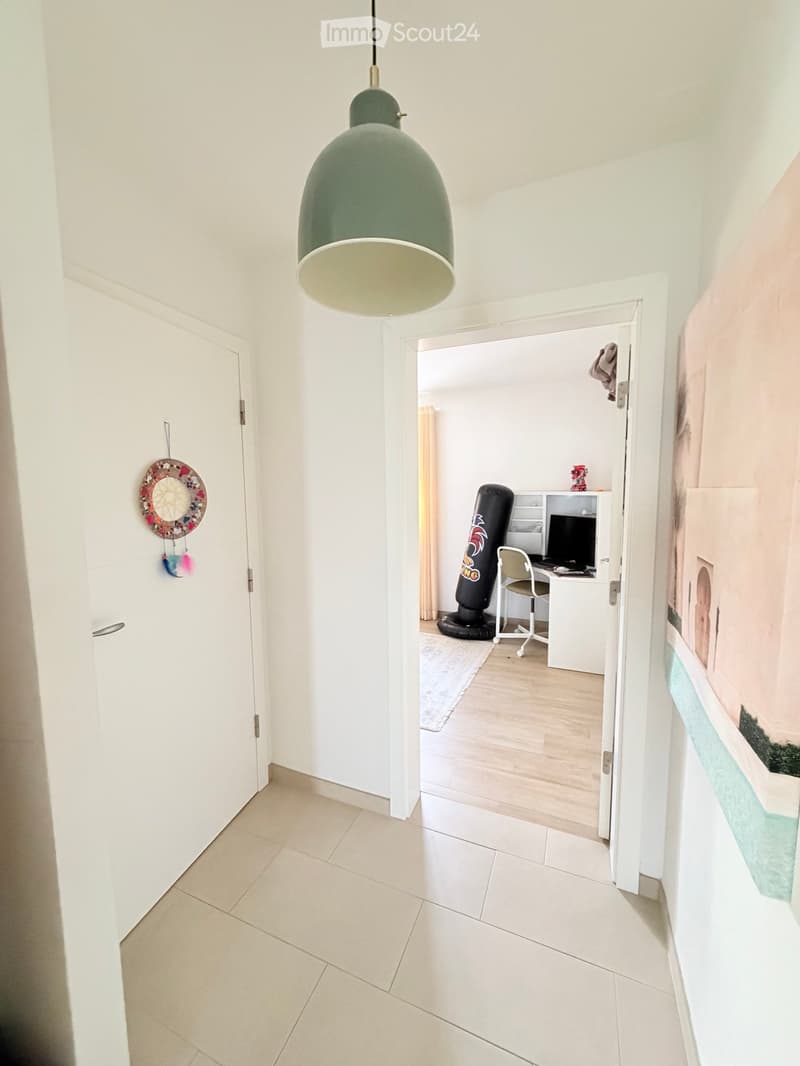 A clean, modern hallway with white walls, tiled flooring, and a green pendant light. An open doorway leads to a furnished room with a desk, chair, computer monitor, and a punching bag.