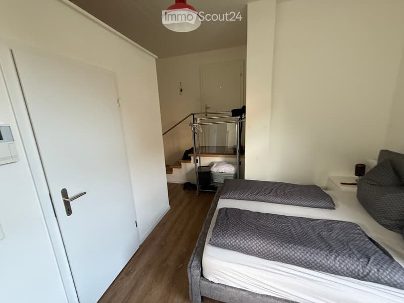 Cozy bedroom with a double bed, nightstand, and lamp. Wooden flooring and white walls. A staircase leads to another level.