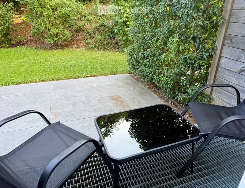 A patio area with a black metal table and two matching chairs, surrounded by greenery and a paved walkway.