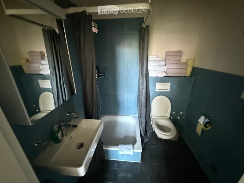 Bathroom with a bathtub, sink, two toilets, and neatly stacked towels on shelves.
