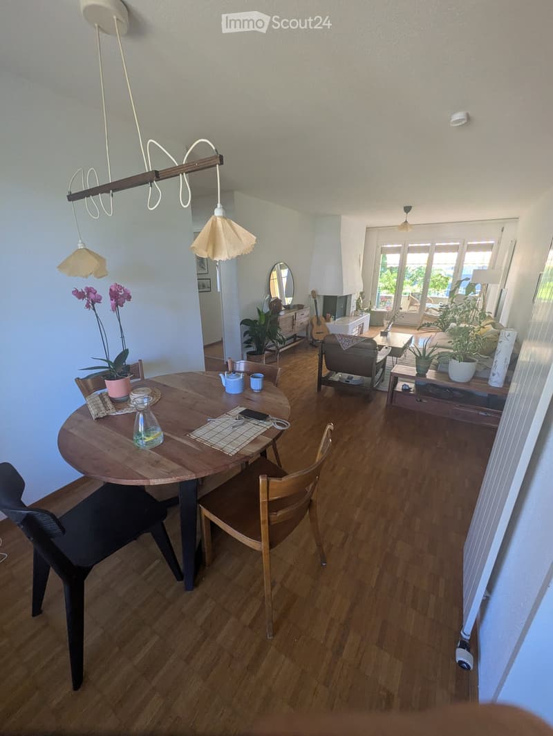 Bright, open dining and living area with wooden flooring, a round wooden dining table with chairs, a sofa, potted plants, a guitar, and a hanging lamp.