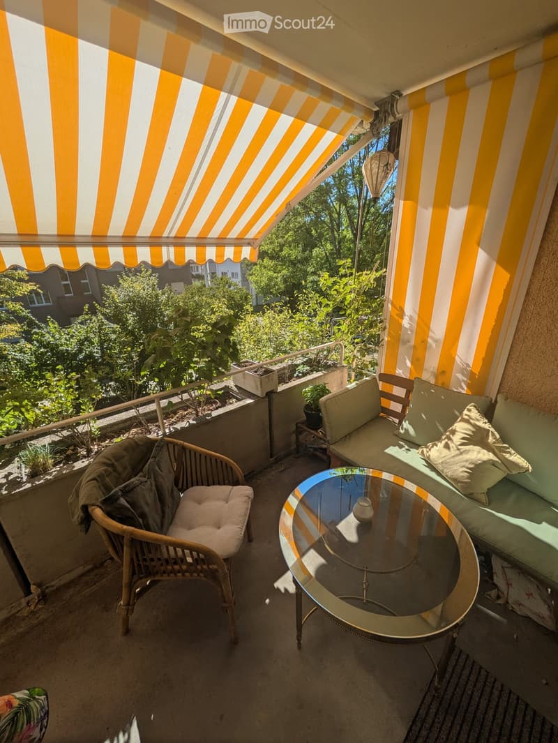 Balcony with striped awning, sofa, wicker chair, glass table, and small potted plants.