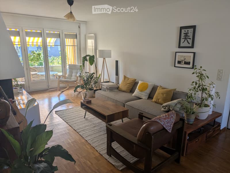 Modern living room with a grey sofa, wooden coffee table, armchair, floor lamp, and potted plants. Large windows and sliding doors lead to a balcony.