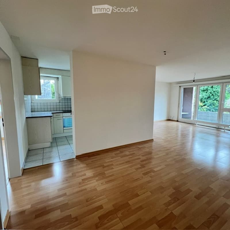 Empty room with wooden flooring, white walls, and large windows leading to a balcony. The adjacent kitchen area is also visible with cabinets and a countertop.