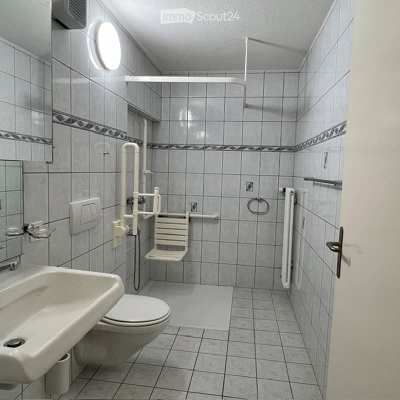 Accessible bathroom with tiled walls and floor, featuring a toilet, sink, shower chair, handrail, and mirror.