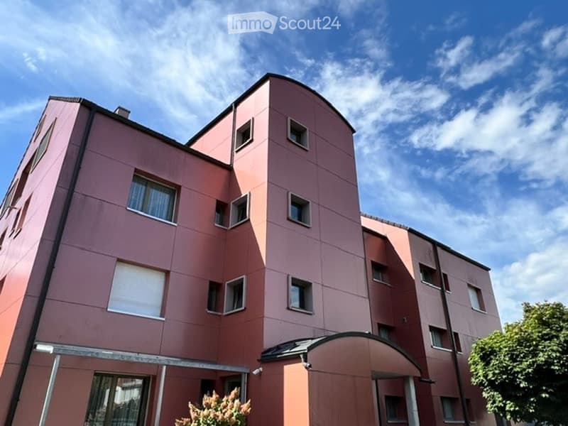 Modern multi-story residential building with pink facade, multiple windows, and a covered entrance area.