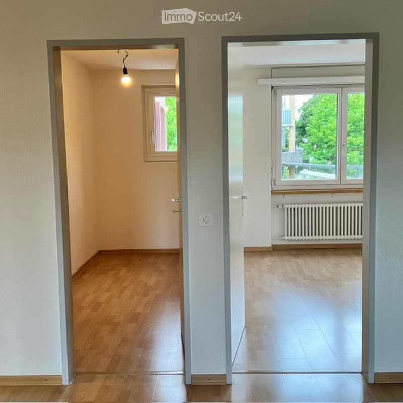 Empty unfurnished room with wooden flooring, white walls, and a window with a view of greenery. The room is connected to another similarly empty space through an open doorway.