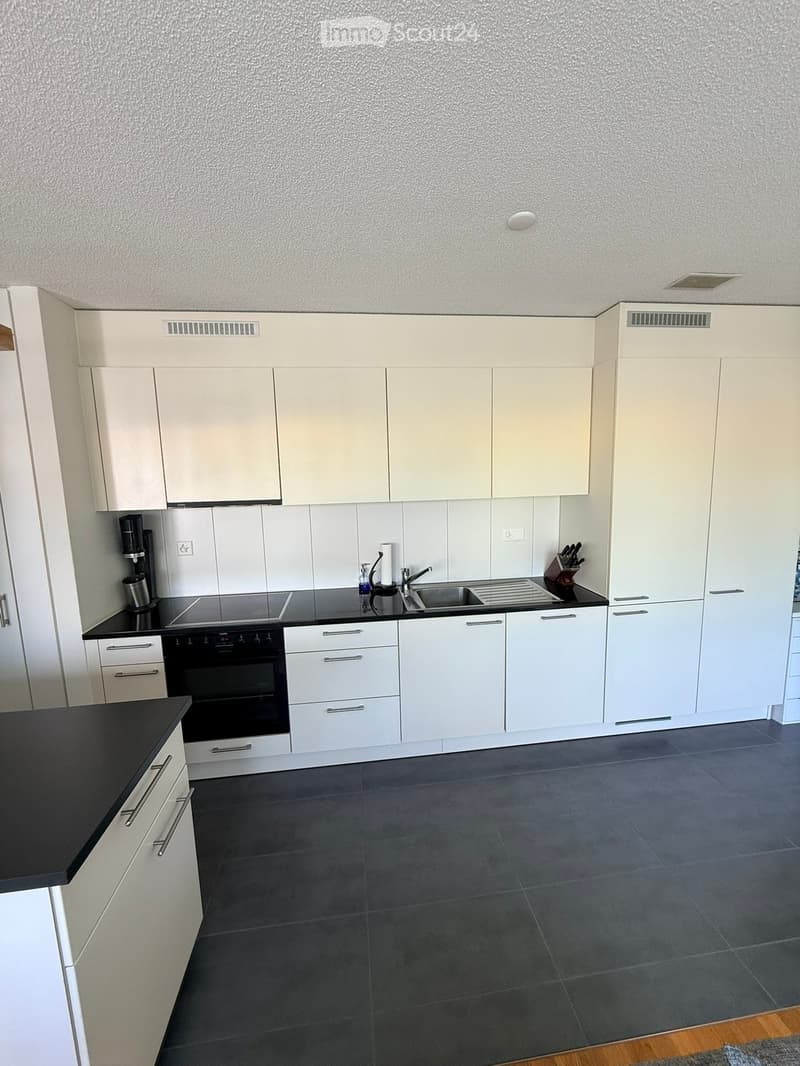 Modern kitchen with white cabinets, black countertops, and grey tiled floor. Features an oven, sink, faucet, coffee maker, and knife block.