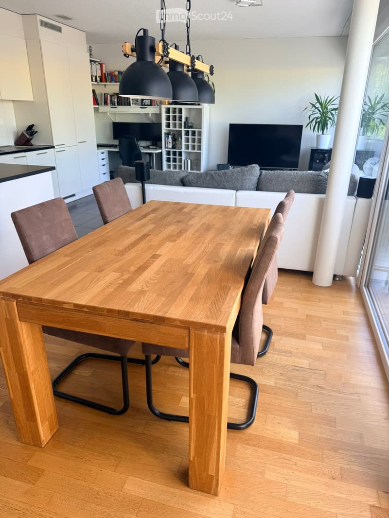 A modern, open-plan living and dining area with a wooden dining table, four chairs, a grey sofa, a television, and a hanging lamp. The space features light wooden flooring and large windows with a view.