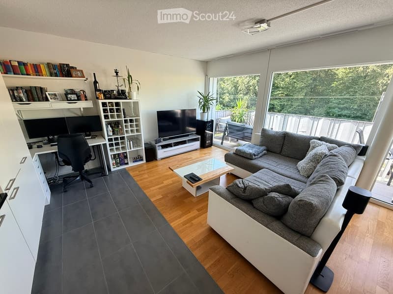 Modern living room with a grey sectional sofa, coffee table, desk with chair, bookshelf, wine rack, television, and plants. Large windows offer a view of greenery.