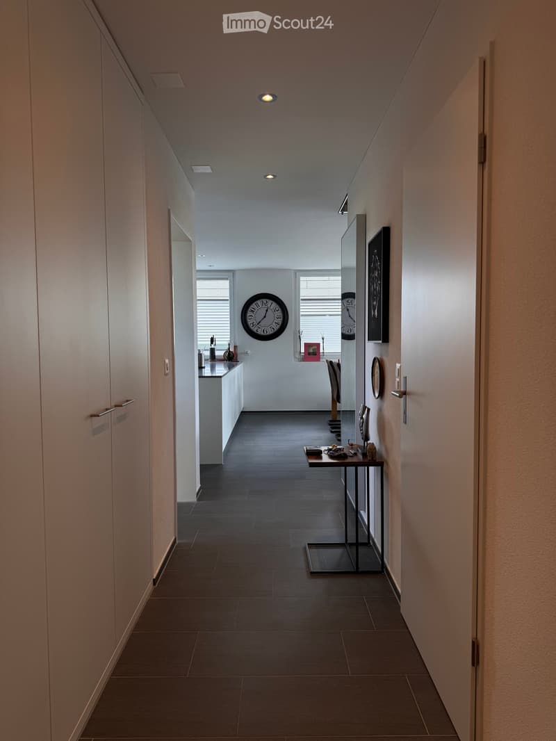 Modern hallway with dark tiled floor, white walls, built-in cabinets, and a small table with decorative items. A large wall clock is visible in the background.