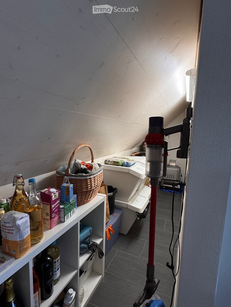 A small, well-lit room with a slanted ceiling, featuring a white shelf holding various items including bottles, a basket, and food products. A vacuum cleaner is mounted on the wall, and a small cabinet is visible in the background.