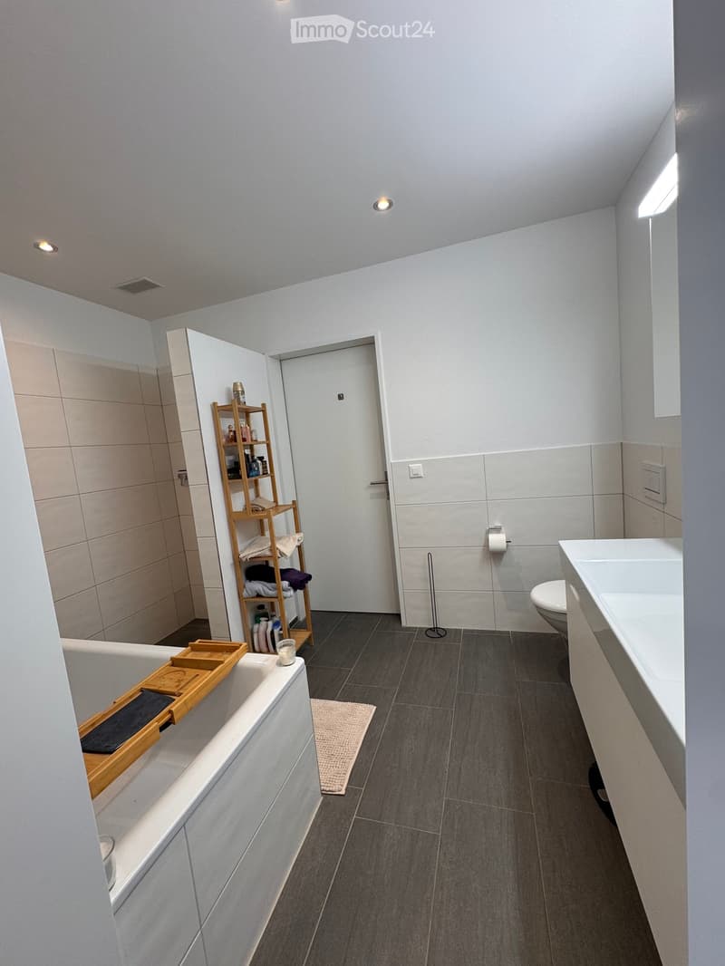 Modern bathroom with a bathtub, toilet, sink, wooden shelf with towels and toiletries, and a small rug on the floor.